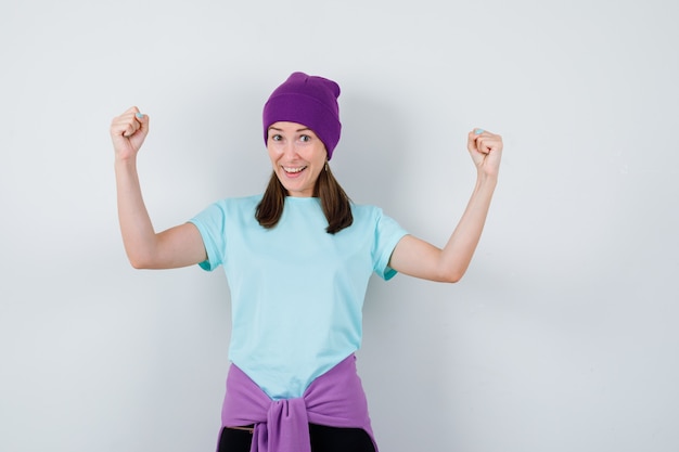 Młoda kobieta w niebieskim t-shirt, fioletowa czapka pokazująca gest zwycięzcy i patrząc na szczęście, widok z przodu.