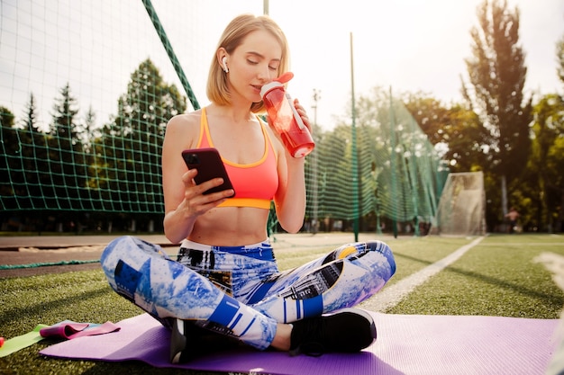 Młoda kobieta w odzieży sportowej siedzi na macie, pije wodę, używa smartfona i odpoczywa po ćwiczeniach sportowych