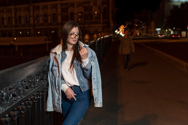 Młoda kobieta w okularach iw swobodnym stylu na wieczornym tle miasta Stylowa dziewczyna spaceruje nocną ulicą miasta