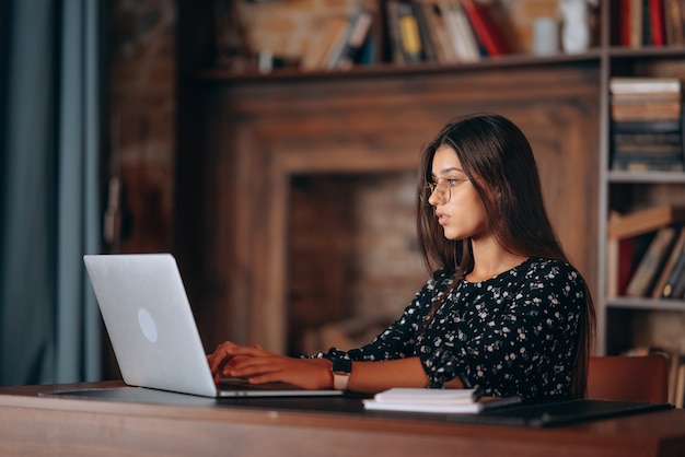 Młoda kobieta w okularach pracuje przy laptopie, siedząc przy stole