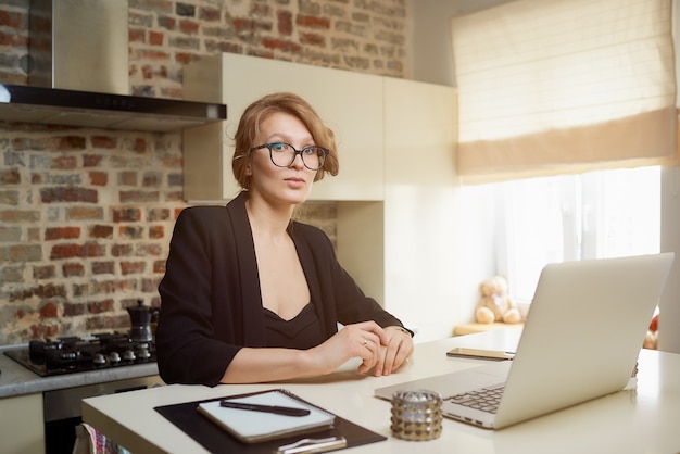 Zdjęcie młoda kobieta w okularach pracuje zdalnie na laptopie w swojej kuchni. blond dziewczyna słucha swoich kolegów na wideokonferencji w domu.