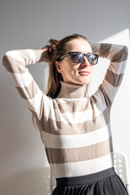 Młoda kobieta w okularach przeciwsłonecznych w pasiastym swetrze na białej ścianie