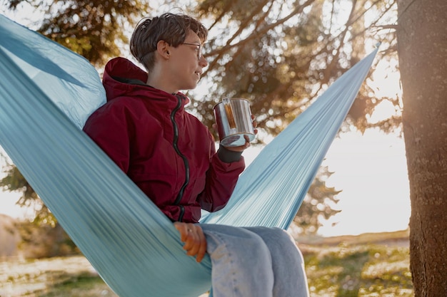 Młoda kobieta w okularach siedzi w hamaku z termosem w lesie i cieszy się rano