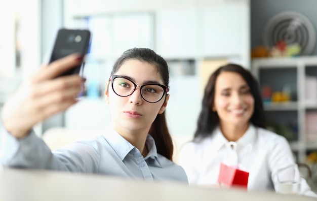 Młoda kobieta w okularach trzyma telefon przed twarzą i uśmiecha się do kolegów w biurze Rozmowa wideo lub selfie w pracy