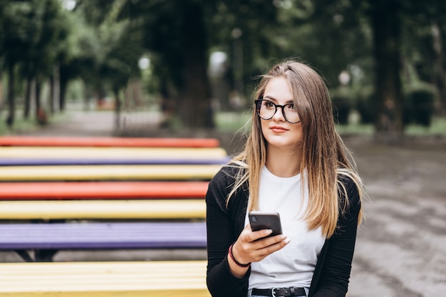 Młoda kobieta w okularach wygląda na telefon. Siedząc na ławce w parku. Pojęcie komunikacji i sieci społecznościowych