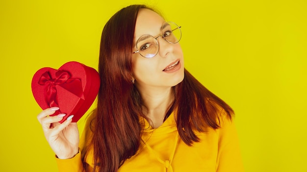 Młoda kobieta w okularach z czerwonym pudełkiem w formie serca na żółtym tle Szczęśliwa kobieta potrząsająca romantycznym prezentem i uśmiechnięta Koncepcja prezentu na walentynki i międzynarodowy dzień kobiet