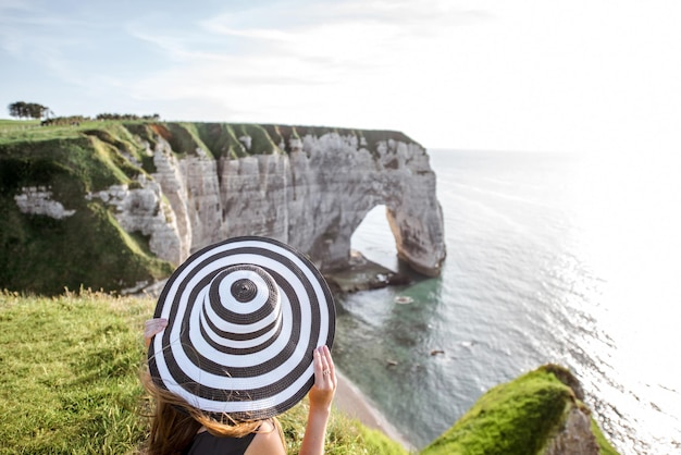 Młoda kobieta w pasiastym kapeluszu, ciesząca się wspaniałym widokiem na słynne skaliste wybrzeże w pobliżu miasta Etretat we Francji