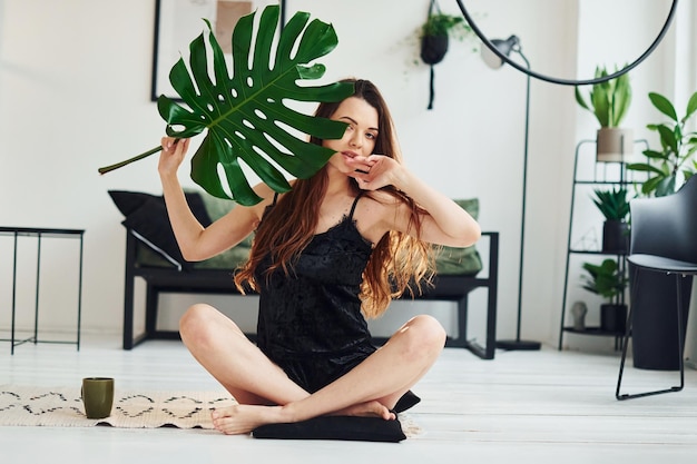 Młoda kobieta w piżamie siedzi na podłodze w pomieszczeniu w ciągu dnia i trzyma duży zielony liść