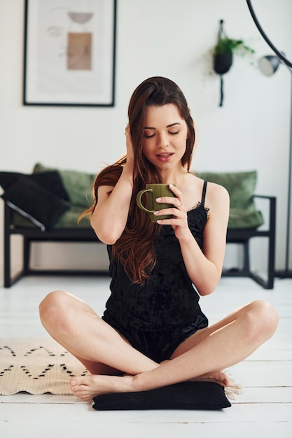 Młoda kobieta w piżamie siedzi na podłodze w pomieszczeniu w ciągu dnia z kubkiem napoju