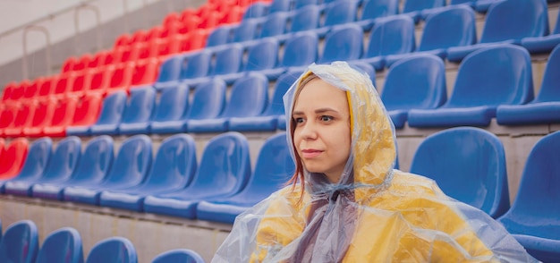 Młoda kobieta w płaszczu siedzi na trybunach stadionu sama w deszczową pogodę Kobieta widz w mokrym płaszczu wodoodpornym na stadionie ulicznym podczas zawodów