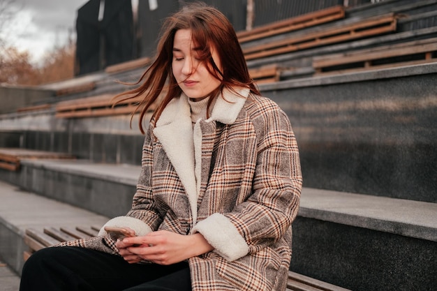 Młoda kobieta w płaszczu ze smartfonem w dłoniach siedzi samotnie na drewnianej ławce na ulicy Praca na urządzeniu mobilnym poza domem na zewnątrz Kobieta freelancer blogerka SMS-y na czacie czytanie