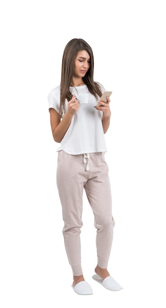 Młoda kobieta w przypadkowych ubraniach trzyma filiżankę kawy i patrzy na ekran swojego smartfona. Odosobniony portret