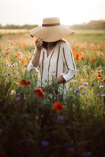 Młoda kobieta w rustykalnej lnianej sukience spacerująca wśród maku i chabrów na letniej łące w świetle zachodzącego słońca Wiejskie powolne życie Cieszące się prostym życiem Dziewczyna w kapeluszu w polnych kwiatach