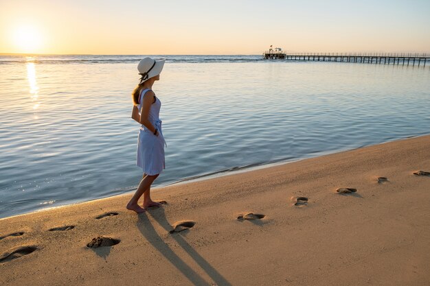 Młoda Kobieta W Słomkowym Kapeluszu I Sukni Chodzi Samotnie Na Pustej Piasek Plaży Przy Zmierzchu Dennym Brzeg.