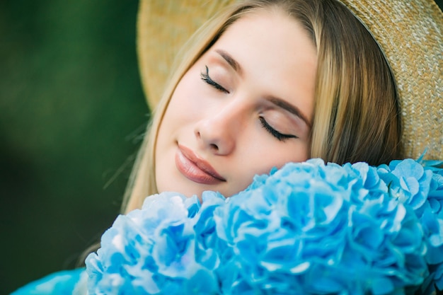 Młoda kobieta w słomkowym kapeluszu pachnie niebieskimi kwiatami hortensji spacerującymi po letniej ulicy