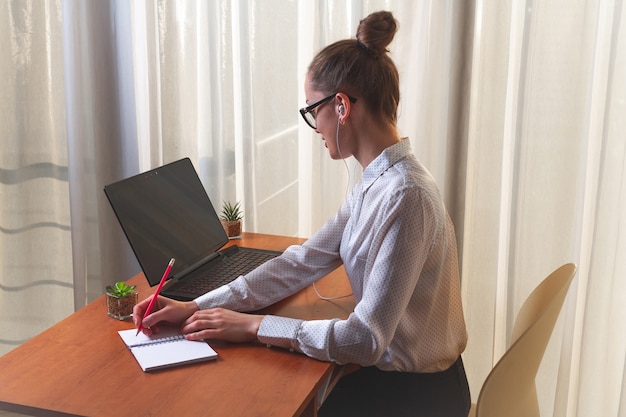 Młoda kobieta w słuchawkach angażowała się w kursy online zdalnie, używając laptopa i zapisując informacje w notatniku