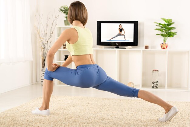 Młoda kobieta w sportowym stroju, atletyczna budowa, sfotografowana od tyłu, ćwiczy w pokoju, przed telewizorem.