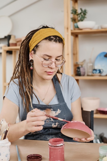 Młoda kobieta w stroju codziennym i okularach malująca glinianą miskę farbą marsala