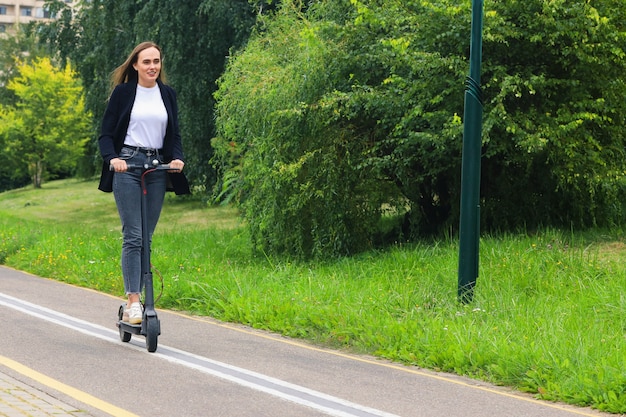 Młoda kobieta w stylowych ubraniach jeździ skuterem elektrycznym po ścieżce rowerowej