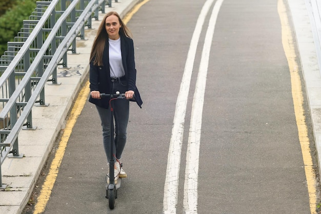 Młoda kobieta w stylowych ubraniach jeździ skuterem elektrycznym po ścieżce rowerowej
