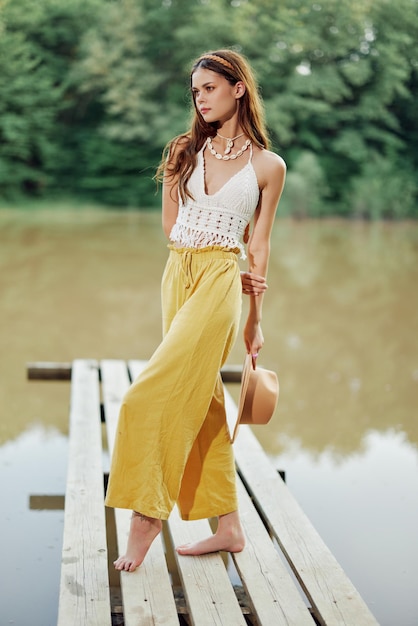Młoda kobieta w stylu hipisa i ekostroju tańczy na świeżym powietrzu nad jeziorem w kapeluszu i żółtych spodniach w letnim zachodzie słońca
