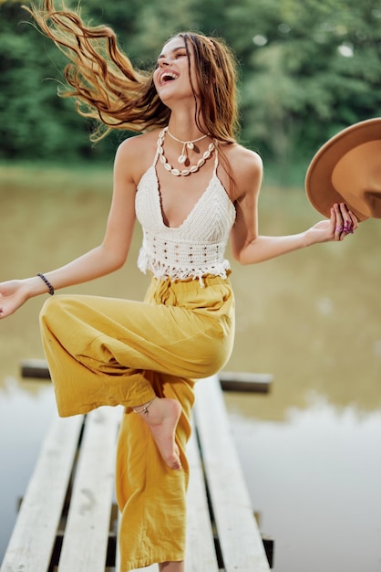 Młoda kobieta w stylu hipisa i ekostroju tańczy na świeżym powietrzu nad jeziorem w kapeluszu i żółtych spodniach w letnim zachodzie słońca