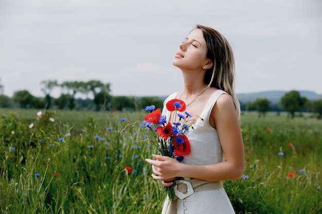 Młoda kobieta w sukience pozostaje w pobliżu pola z bukietem kwiatów Centaurea i maku