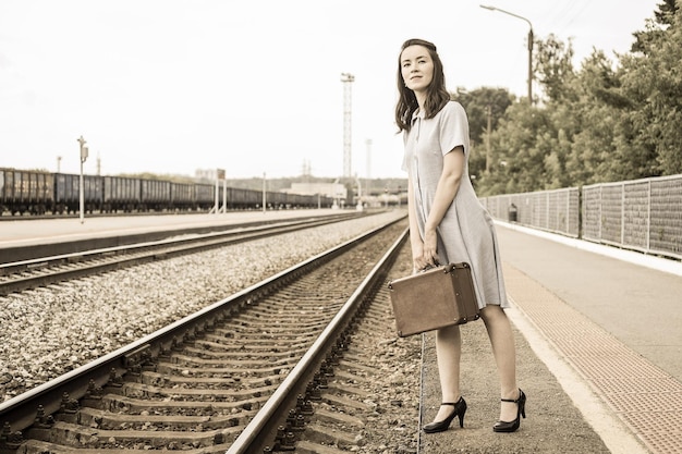 Młoda kobieta w sukience retro z zabytkową walizką w dłoniach stoi na peronie kolejowym i czeka na przyjazd pociągu Kobieta czeka na pociąg