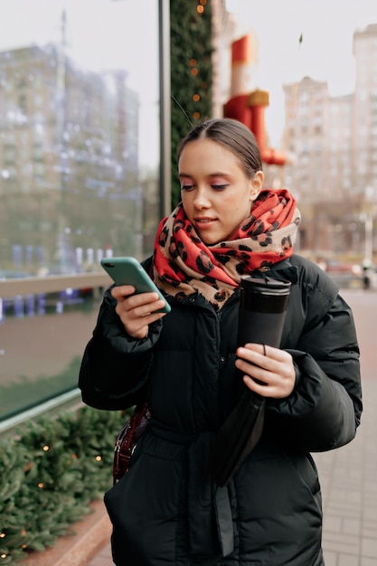 Młoda kobieta w świetnym nastroju śmieje się podczas przewijania smartfona Dziewczyna w ciemnej kurtce pozuje na rozmytym tle miasta