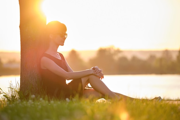 Młoda kobieta w swobodnym stroju siedzi na zielonym trawniku relaksuje się po stronie jeziora w ciepły wieczór Letnie wakacje i koncepcja podróży