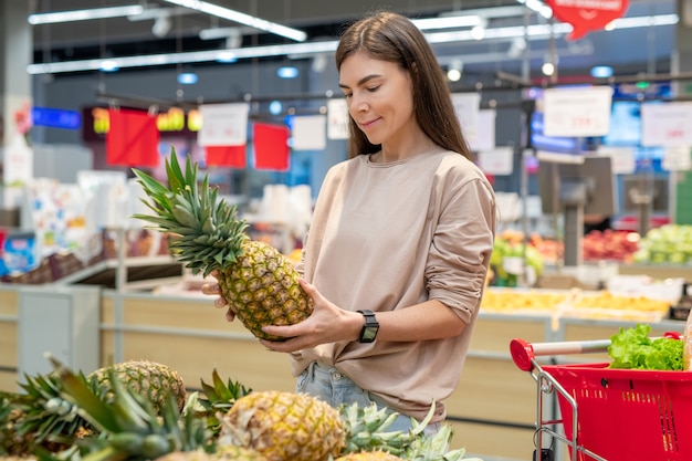 Młoda kobieta w swobodnym stroju, wybierająca najlepszego ananasa w nowoczesnym supermarkecie, kopia przestrzeń