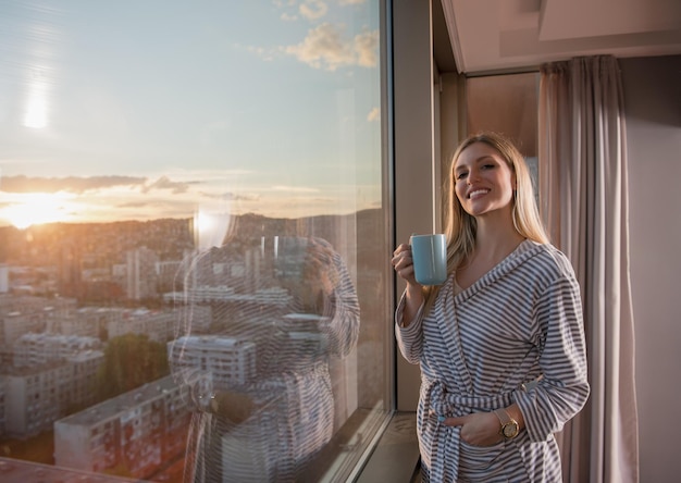 młoda kobieta w szlafroku, ciesząc się wieczorną kawą i pięknym krajobrazem zachodu słońca miasta, stojąc przy oknie