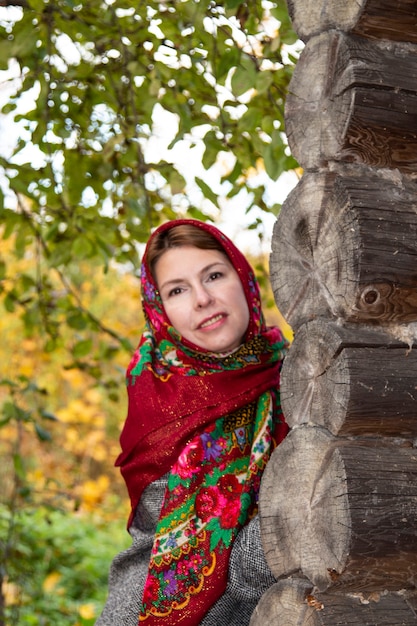 Młoda kobieta w tradycyjnej rosyjskiej chuście rozgląda się za rogiem drewnianego domu. Czerwona chusteczka Pawłowsk Jarosław Rosja 10.10.2019