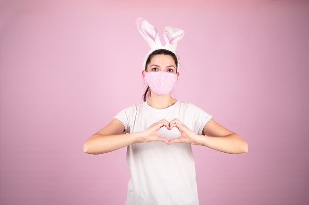 Młoda kobieta w wieku noszenie maski koronawirusa robi kształt symbolu serca rękami na różowym tle. Koncepcja wielkanocna. Wysokiej jakości zdjęcie