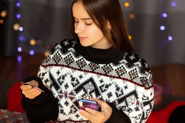 Młoda kobieta w zimowym swetrze robi świąteczne zakupy przez telefon w domu online