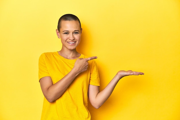 Młoda kobieta w żółtej koszulce z żółtym tłem w studiu podekscytowana, trzymając kopię przestrzeni na dłoni