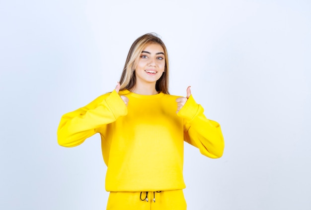 Młoda kobieta w żółtym dresie daje aprobaty na białej ścianie