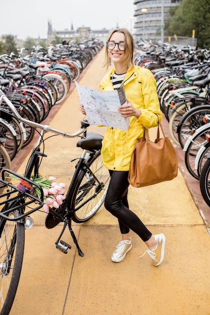 Młoda kobieta w żółtym płaszczu przeciwdeszczowym stojąca z mapą turystyczną na parkingu rowerowym w Amsterdamie