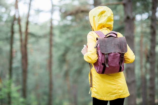 Młoda kobieta w żółtym płaszczu przeciwdeszczowym stojąca z plecakiem w zielonym lesie sosnowym