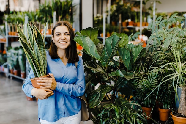 Młoda kobieta wybiera rośliny domowe i doniczki w sklepie lub centrum ogrodniczym Pani trzyma w dłoniach dużą Sansevieria patrzy w kamerę