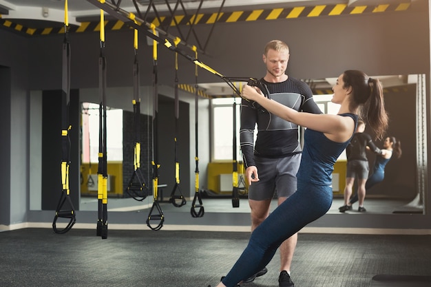 Młoda kobieta wykonując trening TRX w siłowni z osobistym trenerem. Sportowa dziewczyna robi ćwiczenia rozciągające z elastyczną liną, kopia przestrzeń