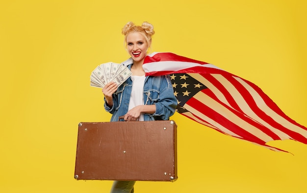 Młoda kobieta z amerykańską flagą trzyma w rękach walizkę podróżną i banknoty pieniędzy. Zaskoczona wesoła twarz.