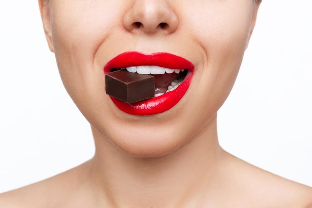 Młoda kobieta z błyszczącymi czerwonymi ustami, trzymająca w zębach gorzki czekoladowy cukierek