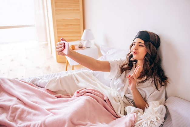 Młoda kobieta z ciemnym włosy bierze selfie pozuje na telefonu kamerze. Sam w sypialni. Piękny model w piżamie. Poranne światło dzienne.