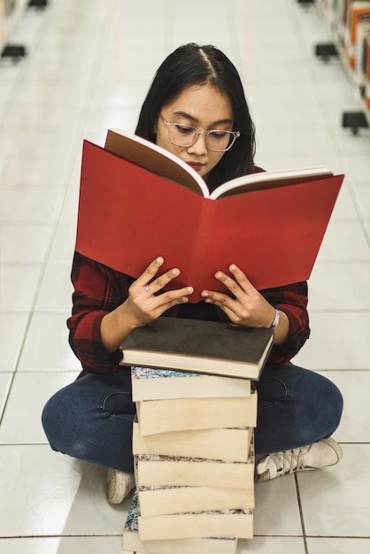 Młoda kobieta z college'u siedzi ze skrzyżowanymi nogami na podłodze między bibliotecznymi półkami i czyta