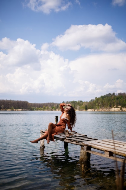 Młoda kobieta z długie falowane kręcone włosy w długiej sukni z gipiury boso latem w lesie nad jeziorem o zachodzie słońca stojąc na pantone na drewnianym moście. Letni słoneczny dzień