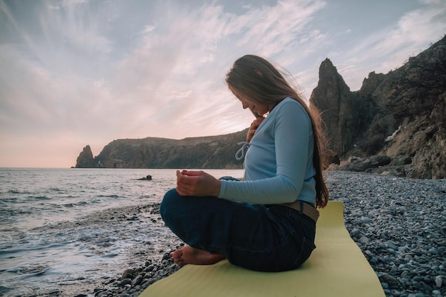 Młoda kobieta z długimi włosami ćwiczy rozciąganie na świeżym powietrzu na macie do jogi nad morzem w ciepły zachód słońca