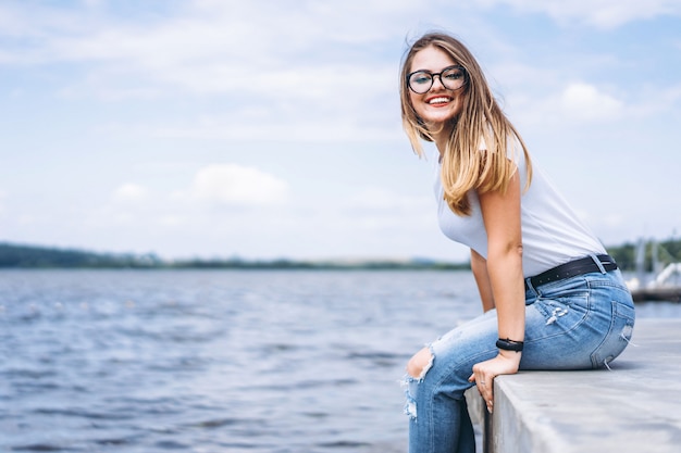 Młoda kobieta z długimi włosami w stylowe okulary pozowanie na betonowym brzegu w pobliżu jeziora. Dziewczyna ubrana w dżinsy i koszulkę, uśmiechając się i patrząc