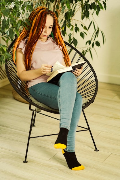 Młoda kobieta z dredami czytająca książkę siedząca na krześle w otoczeniu roślin doniczkowych