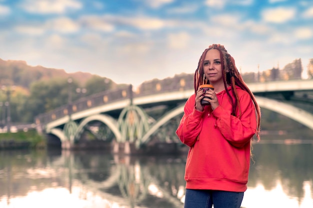 Młoda kobieta z dredami trzymająca filiżankę kawy stojącą w pobliżu jeziora rzeki na tle dużego mostu w parku miejskim